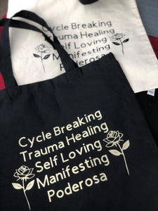 Black tote laying on top of a natural canvas tote. Aerial view image. Black tote has gold artwork which reads CYcle Breaking, Trauma Healing, Self Loving, Manifesting, Poderosa centered between two elegant gold roses.